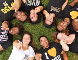 Students laying on grass in a circle facing the camera and pointing up